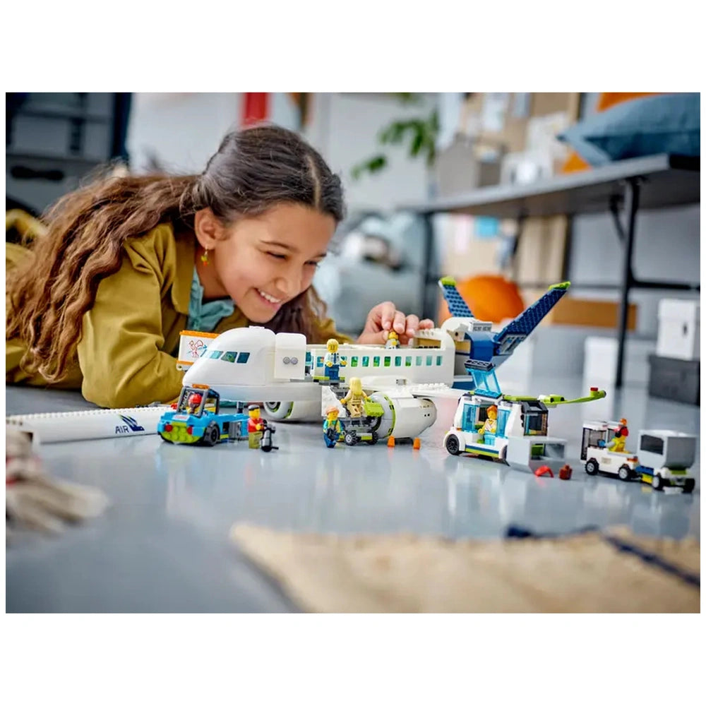 A young girl with curly hair smiles while playing with a LEGO® City Passenger Airplane set, surrounded by detailed minifigures and vehicles.
