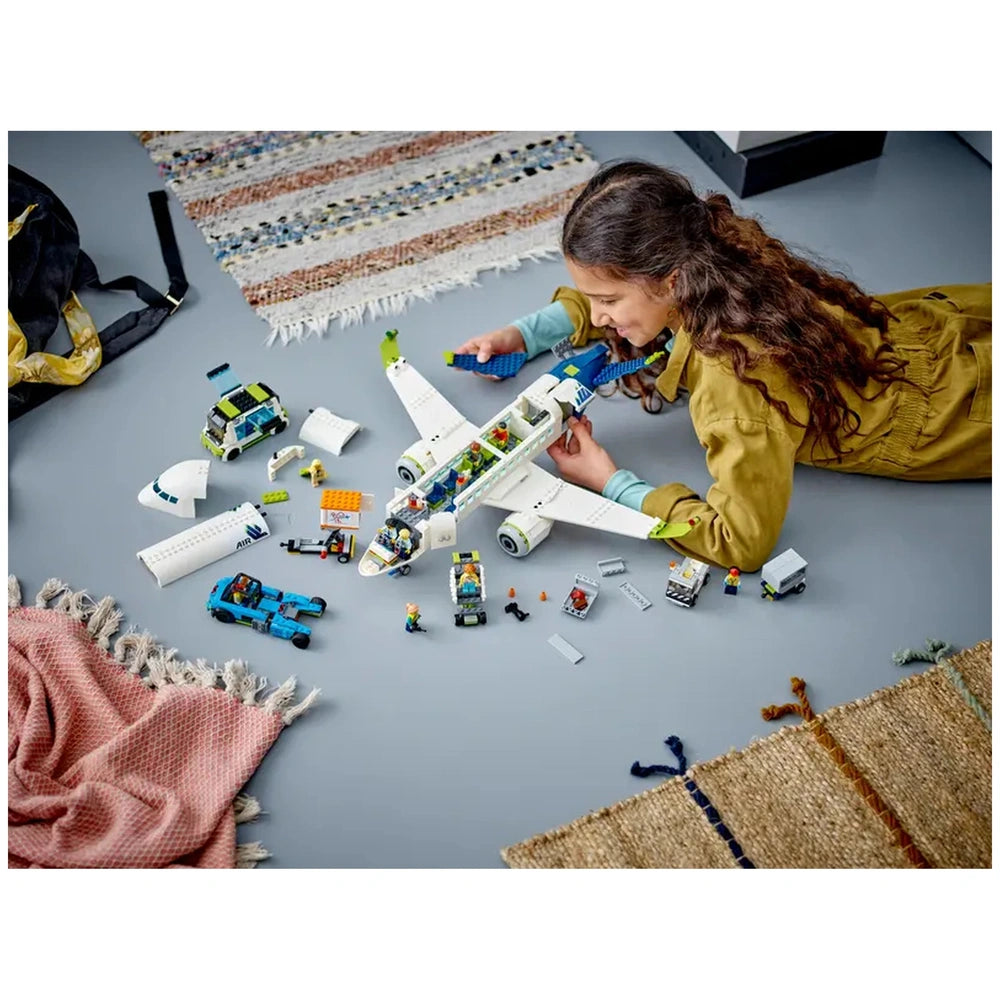 A child with curly hair is lying on a gray floor, assembling a LEGO® City Passenger Airplane. Surrounding them are various LEGO pieces, including vehicles and nine minifigures. A colorful woven rug and fringed fabric are visible in the background, creating a playful and creative atmosphere.