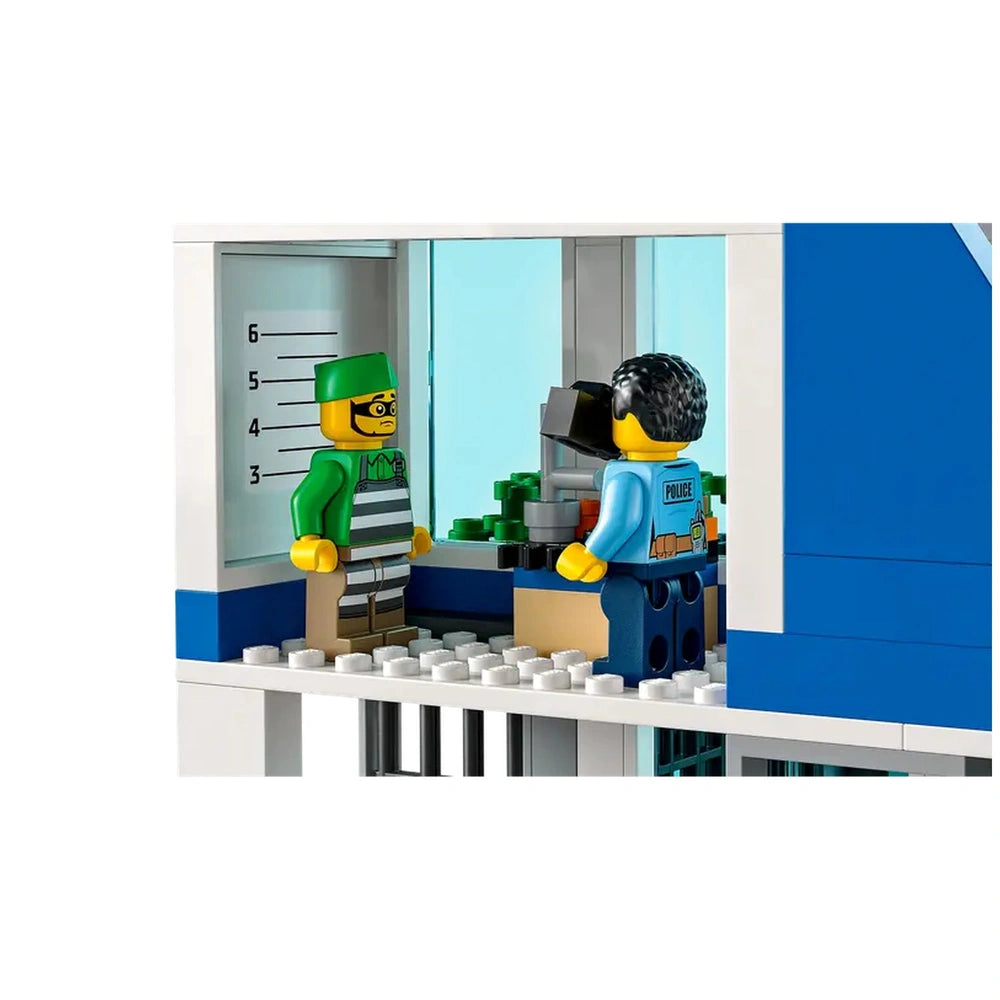 Two LEGO minifigures stand in a police station scene. One is a police officer in a blue uniform with a camera, while the other is a green-capped crook in stripes, standing against a height chart. Behind them, a window reveals a small display of colorful toy items, suggesting an engaging play scenario.