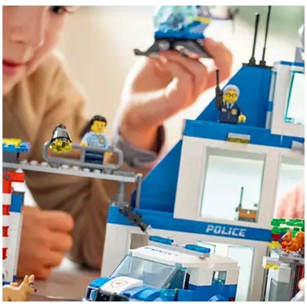 A child builds with the LEGO® City Police Station set, featuring colorful blocks and minifigures. The police station has a detailed structure with windows and a rooftop antenna. A boy holds a helicopter above while another figure stands on a balcony, alongside a patrol vehicle in the foreground.