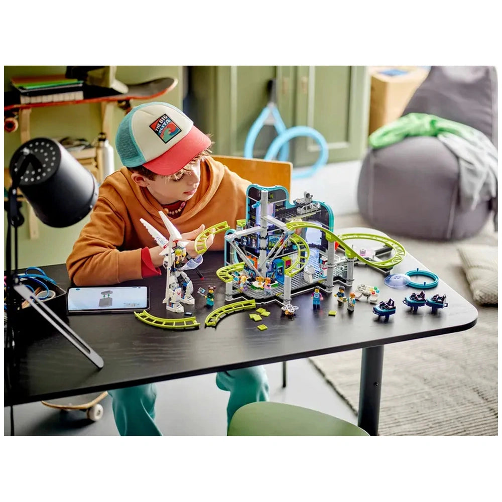 A child with a red and blue cap sits at a black table, focused on a colorful LEGO® City Robot World Roller-Coaster Park set, featuring tracks, a futuristic arcade, and playful characters. A smartphone lies beside them, and the background includes soft furnishings.