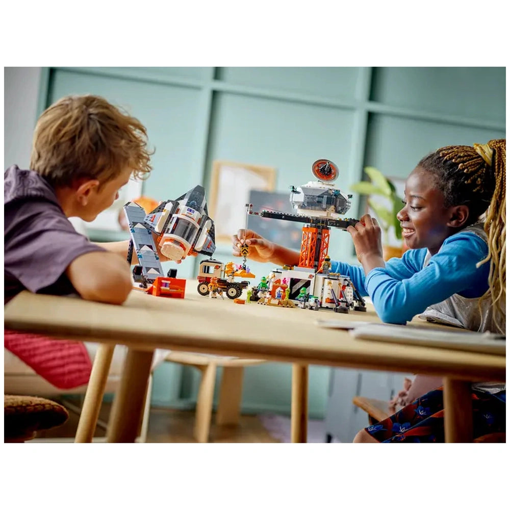 Two children play with the LEGO® City Space Base and Rocket Launchpad set on a wooden table, one holding a spaceship, the other manipulating a crane above the space station. Bright colors and detailed mini-figures enhance their imaginative play in a casual indoor setting.