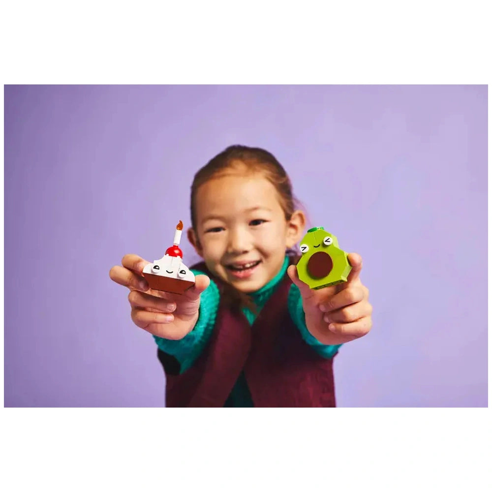 A smiling child holds up a playful cupcake with a candle and a cheerful avocado, set against a purple background.