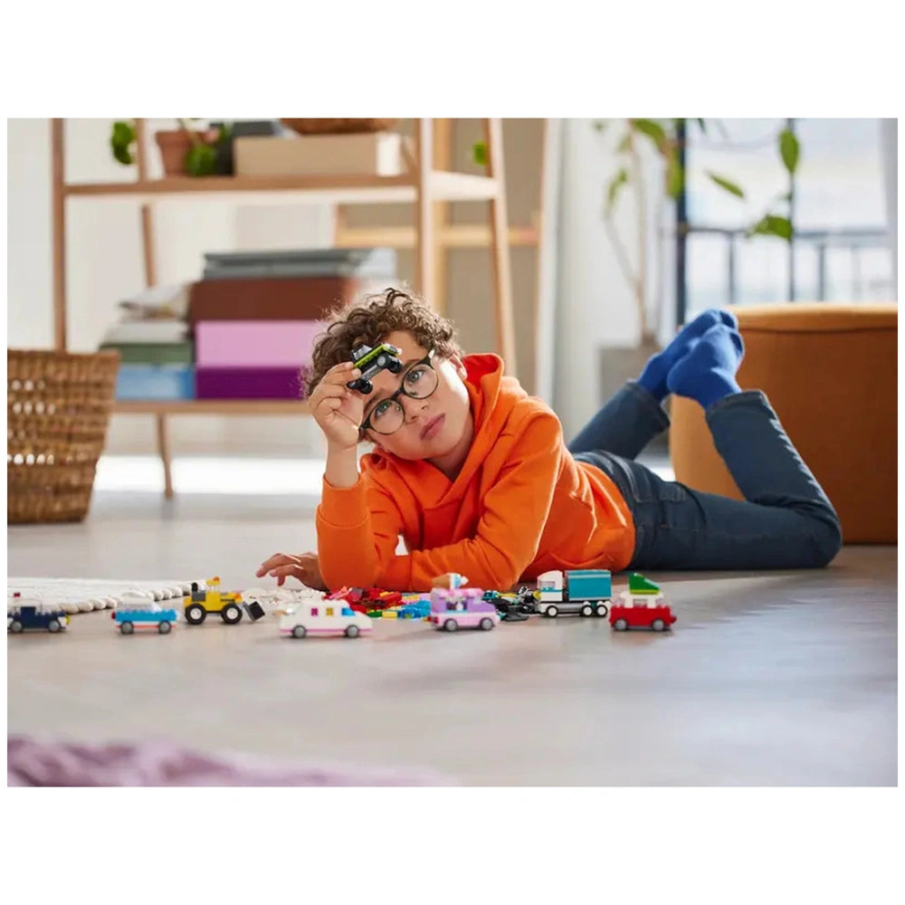 A young boy wearing a bright orange hoodie and glasses lies on the floor, thoughtfully holding a small black vehicle. Surrounding him are various colorful LEGO vehicles, including a pink car and a blue truck, with additional bricks scattered nearby. A cozy, modern room with plants and shelves is visible in the background.