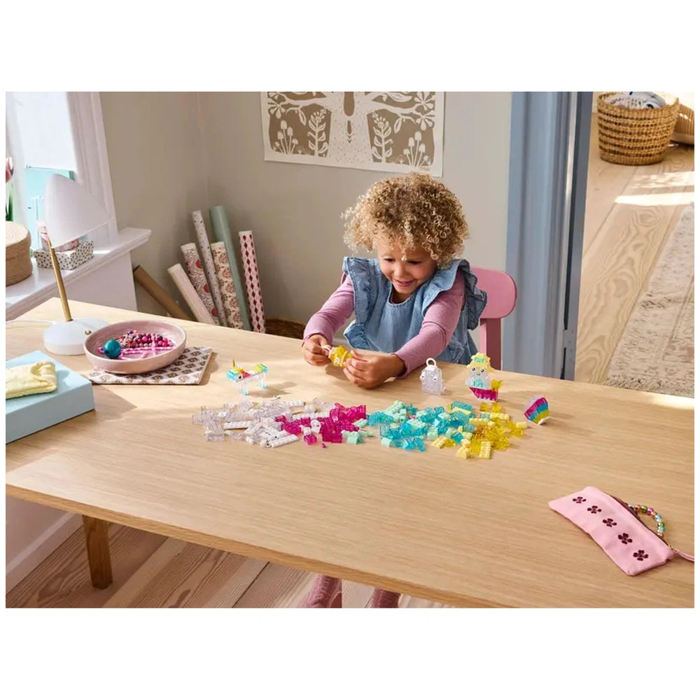 A cheerful child with curly hair sits at a light wooden table, focused on building with colorful transparent LEGO bricks. Surrounding them are a vibrant mix of pink, blue, and yellow pieces, alongside a bowl filled with small gems. A cozy, well-lit room features patterned rolls of craft paper and a decorative wall.
