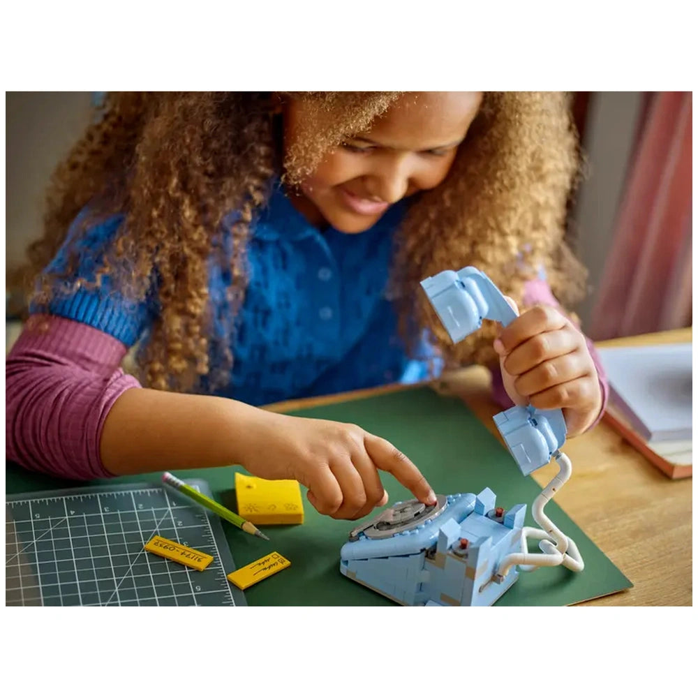 A young girl with curly hair interacts with a blue LEGO Creator 3in1 Retro Telephone set on a green mat. She is lifting the receiver with one hand while pointing at the rotary dial with the other. Nearby, a yellow pencil and sticky notes add color to the workspace.