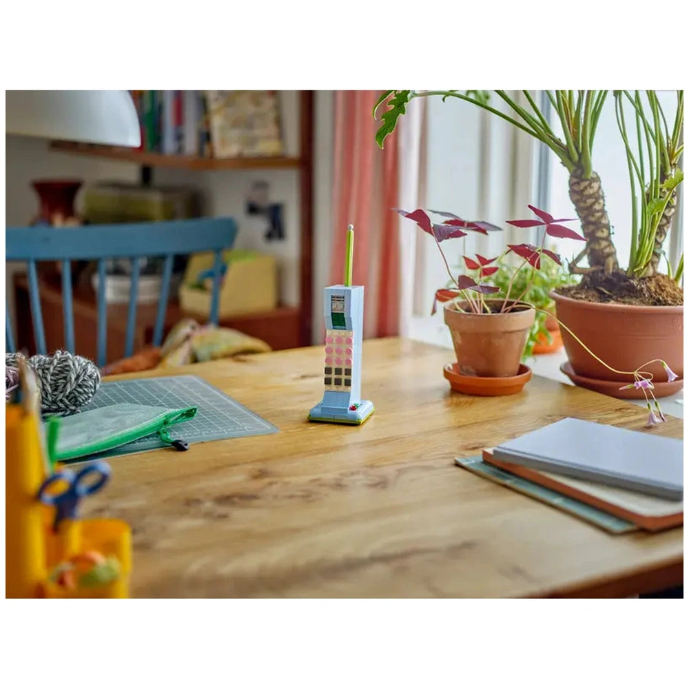 Retro telephone construction set in soft pastel colors, standing on a wooden table with a green sketch pad, potted plants, and craft supplies nearby.