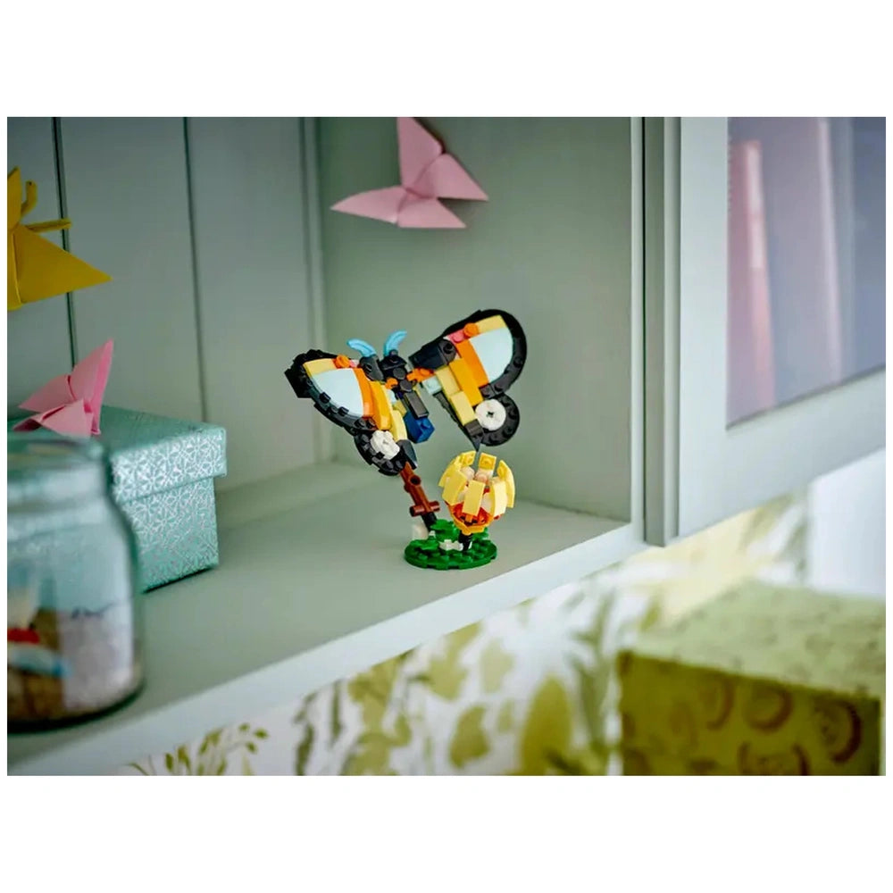 Colorful LEGO butterfly model with movable wings, displayed on a shelf beside a wrapped gift and a glass jar, against a soft green backdrop.