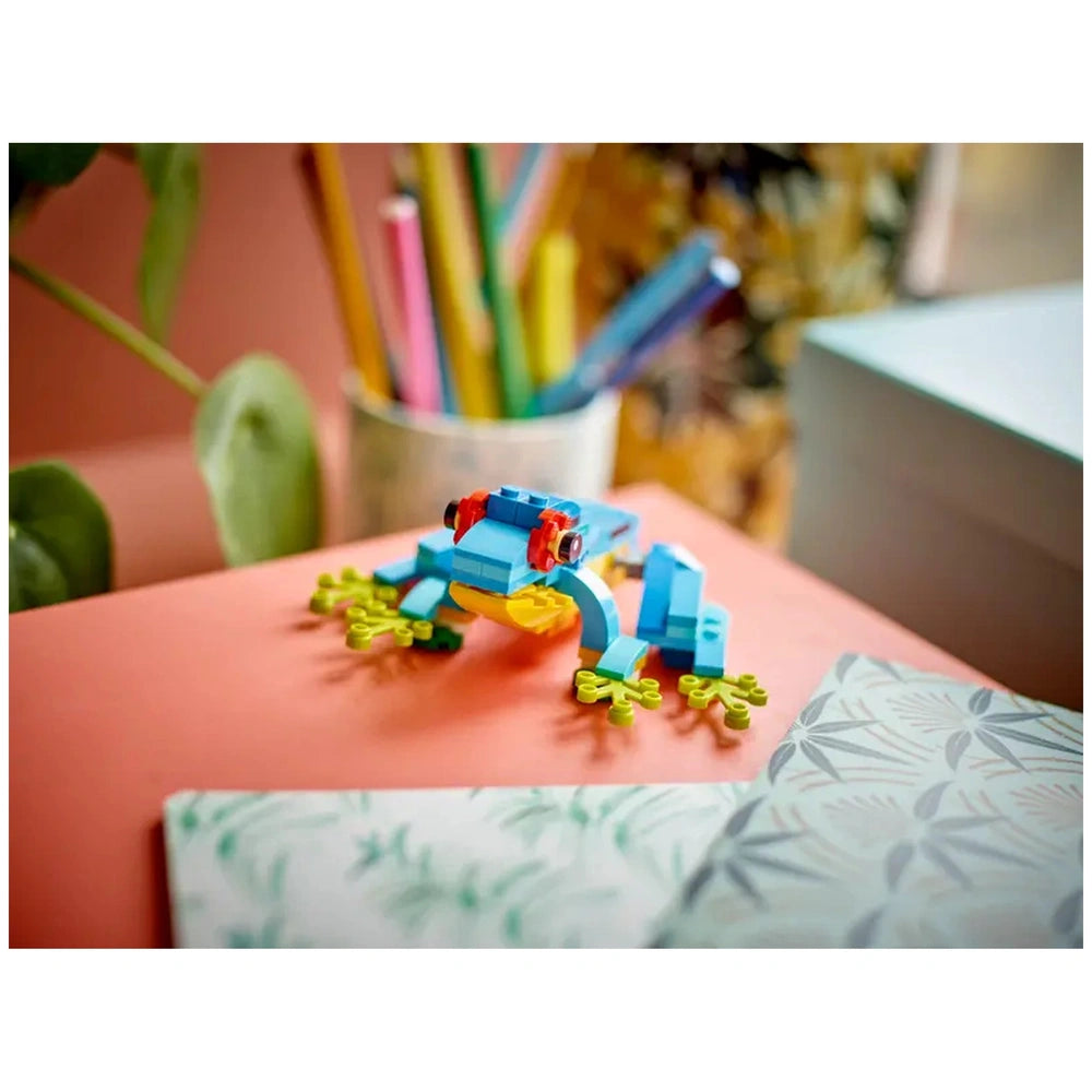 Colorful LEGO® Creator frog in blue and green, with a yellow belly, sitting on a vibrant coral surface, surrounded by plants and colored pencils.