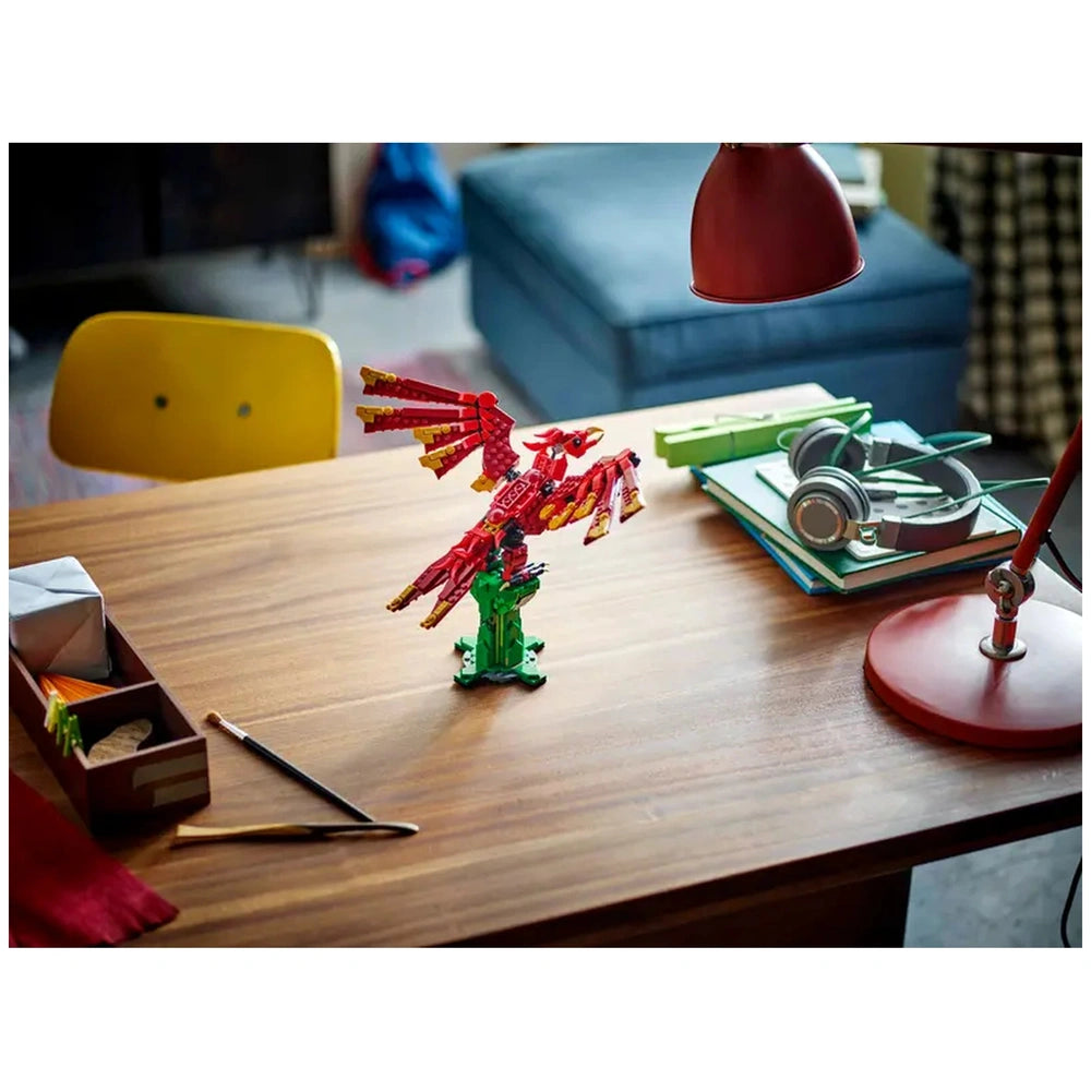 Colorful LEGO® Creator phoenix model stands on a wooden table surrounded by stationery, a lamp, and books in a cozy room setting.