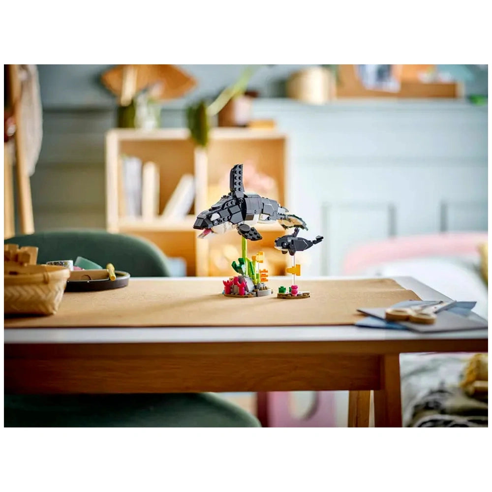 A LEGO Creator set displaying a black and white orca and its calf soaring above a colorful underwater scene with plants and coral on a table, set in a cozy room.
