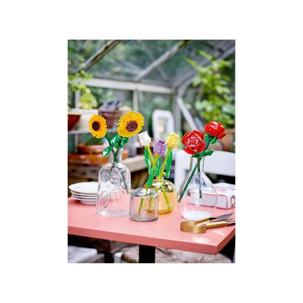 Colorful LEGO flowers in glass vases, featuring yellow sunflowers and red roses, displayed on a pink table in a bright greenhouse.