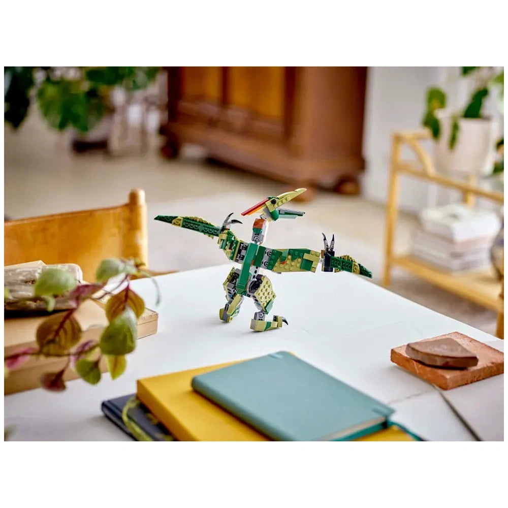 Bright green LEGO Creator dinosaur model stands on a white table, showcasing dynamic poses with extended wings. Nearby are stacked notebooks in yellow and green, and a small decorative piece. A leafy plant and wooden furniture are blurred in the background.