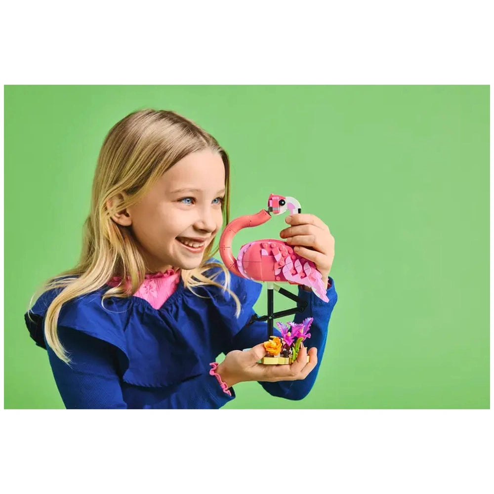 A smiling child holds a LEGO® Creator 3in1 Wild Animals pink flamingo, standing on a plant-covered base, against a bright green background.