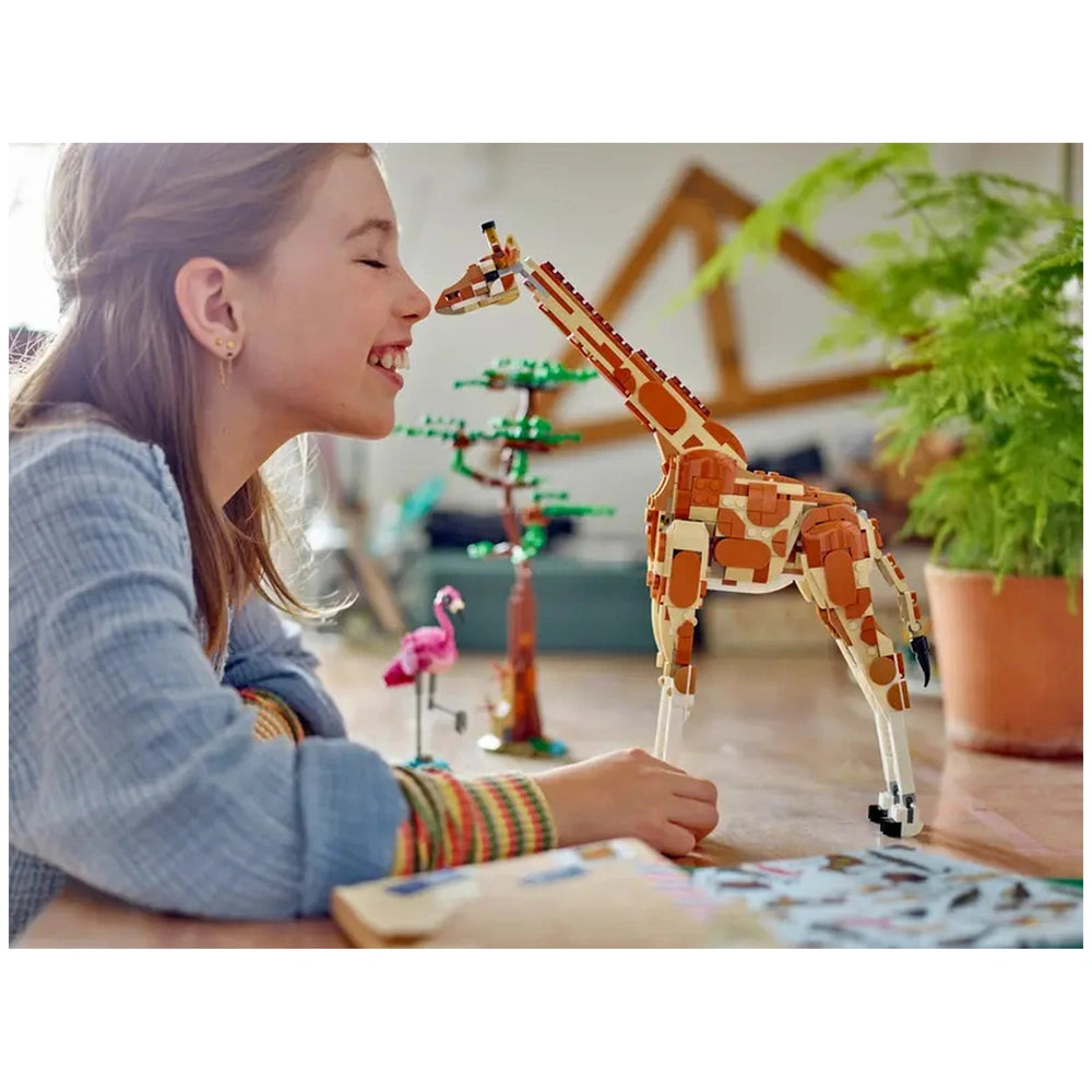 A smiling girl leans towards a colorful LEGO giraffe, showcasing its articulated legs and long neck. A flamingo and a tree are in the background.