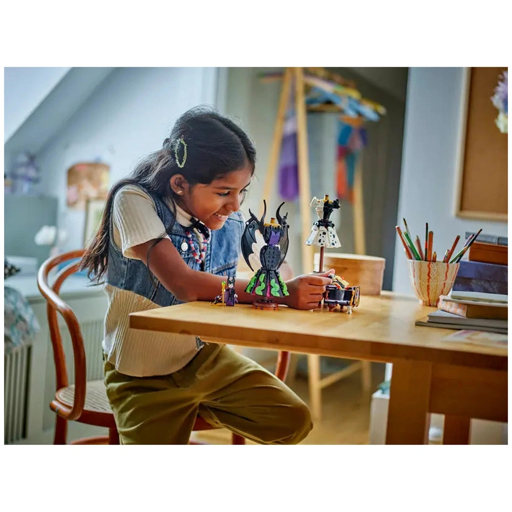 A child smiles while building LEGO® figures of Maleficent and Cruella De Vil on a wooden table, surrounded by art supplies.