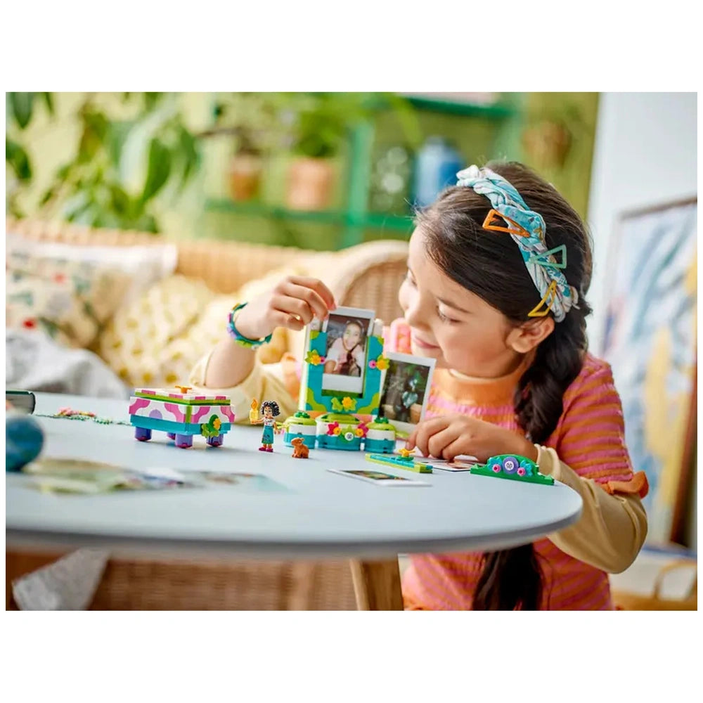 A child joyfully holds two printed photos next to a colorful LEGO® jewelry box and photo frame, surrounded by various small figures and accessories on a light blue table. Bright green plants and a cozy chair add a warm atmosphere to the playful scene.