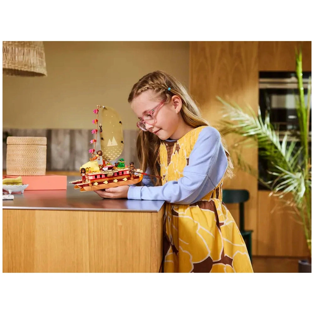 A young girl with glasses admires LEGO® Disney Moana's Adventure Canoe, featuring a vibrant sail and playful design, while seated at a wooden table.