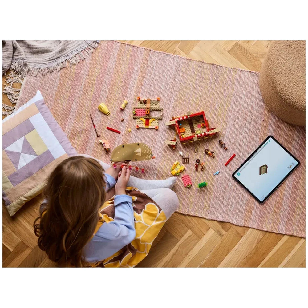 A child sits on a pink rug, assembling LEGO® pieces to create Moana's Adventure Canoe. Colorful bricks are scattered around, alongside a tablet displaying the building model. A cozy pillow sits nearby.