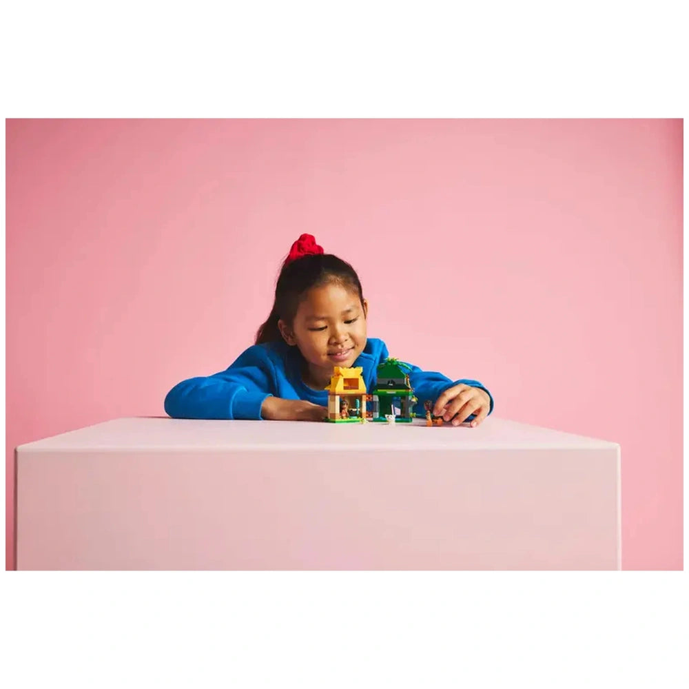 A young child in a blue sweater plays with a LEGO® Disney Moana's Island Fun set, featuring a yellow and green building, against a pink background.