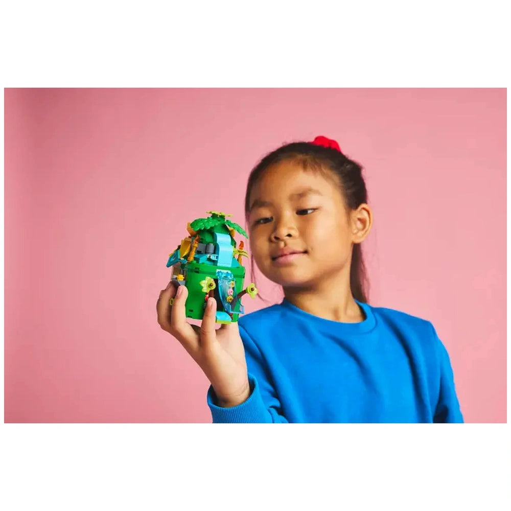 A girl in a blue sweatshirt confidently holds a colorful LEGO® Moana construction set, featuring a vibrant island model with greenery, a slide, and playful details, against a pink background.