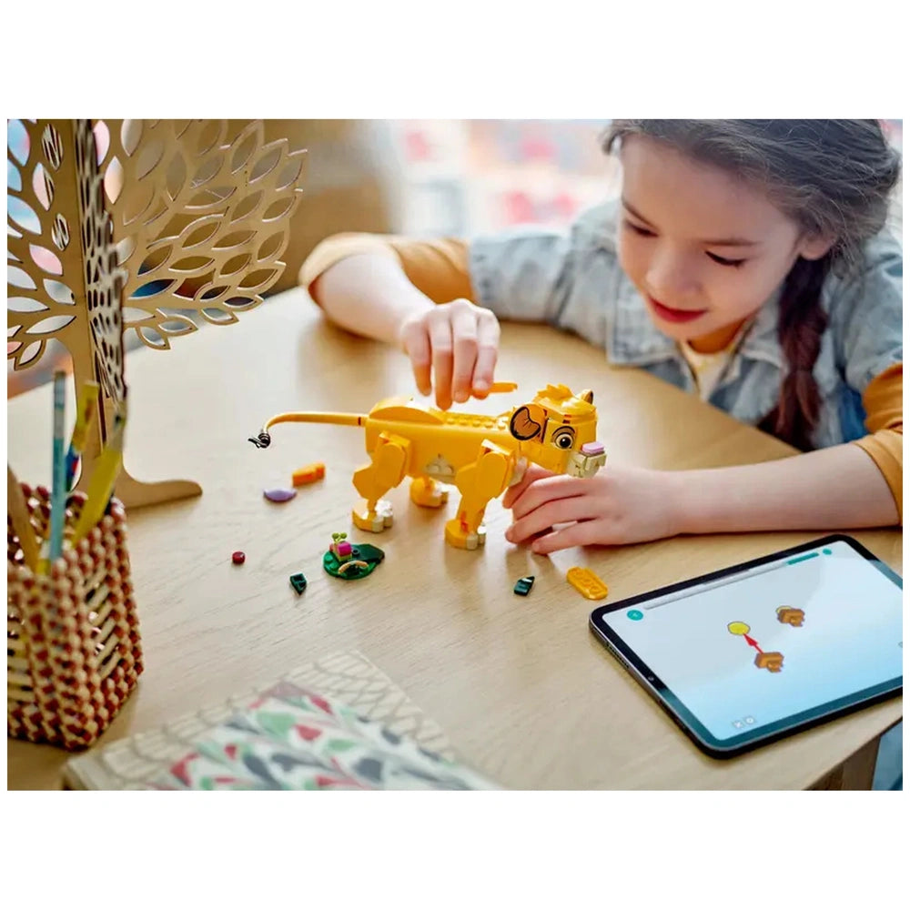 A child creates with the LEGO® Disney Simba the Lion King Cub set, featuring a vibrant yellow Simba figure, on a wooden table. Nearby, a digital tablet displays a drawing of Simba, while colorful building pieces and a playful leaf build are scattered around. A decorative tree sculpture is visible in the background.