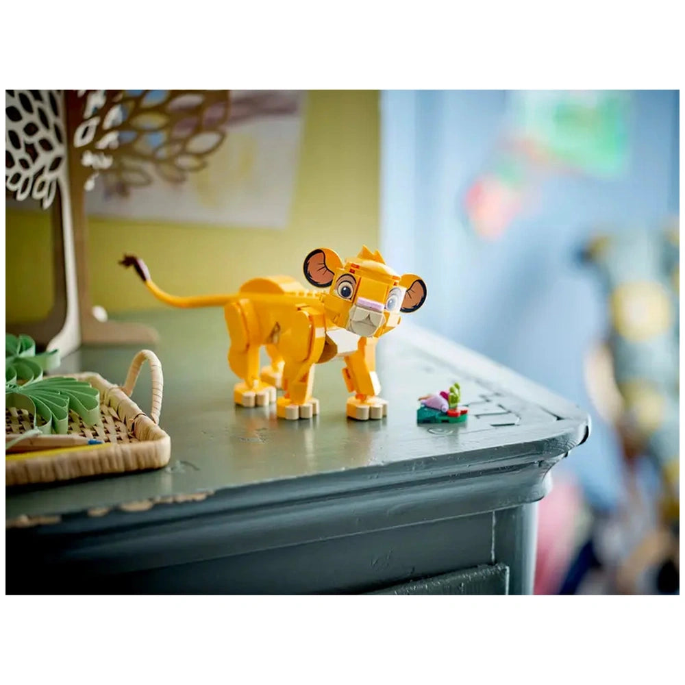 LEGO® Disney Simba the Lion King Cub construction set on a wooden table, featuring a vibrant yellow Simba figure with movable parts and a small leaf build with four colorful 'bugs' beside it.