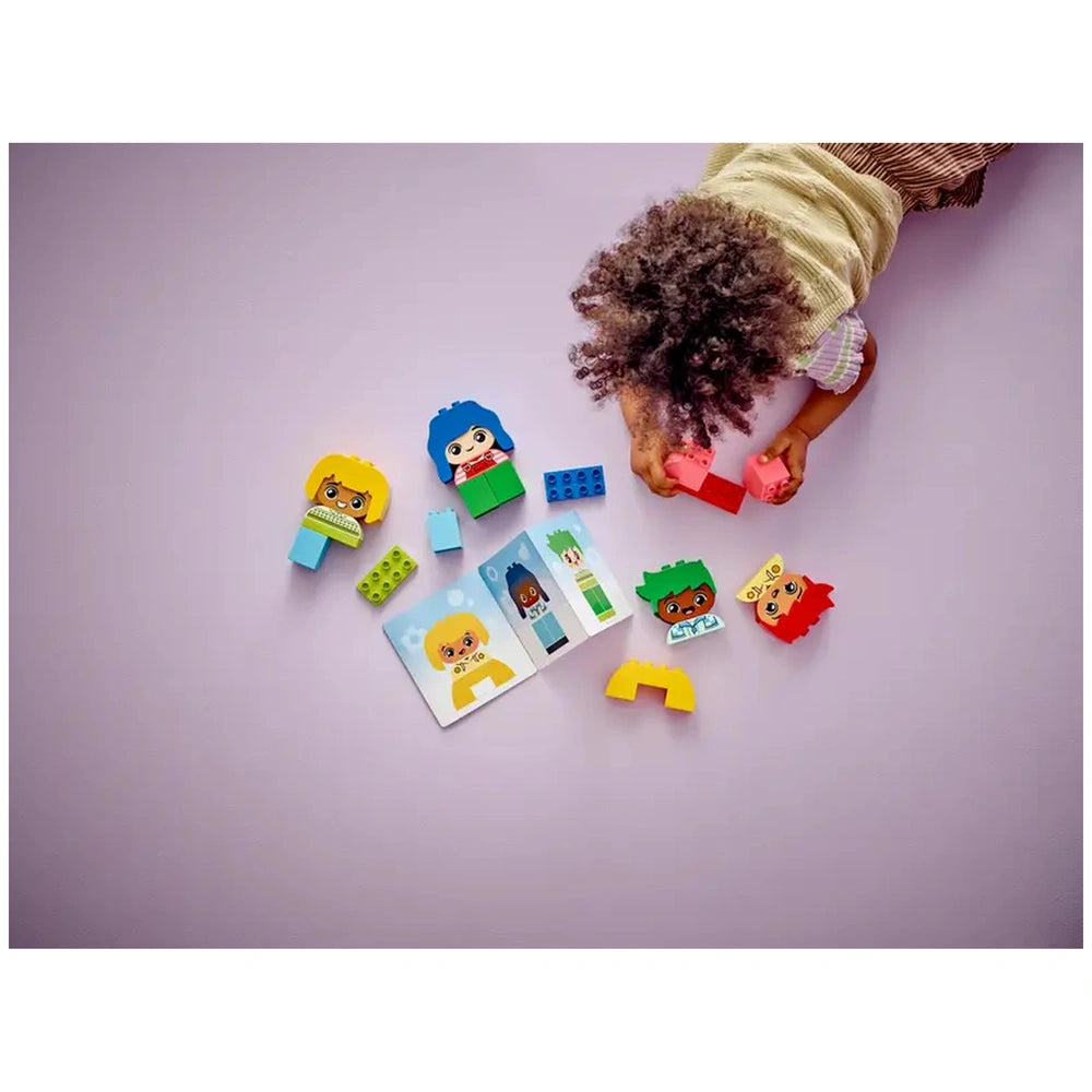 A child with curly hair plays on a purple surface, assembling colorful LEGO® DUPLO® bricks. Visible are character blocks in bright colors, two printed emotion cards, and additional building pieces, promoting emotional learning and creativity.