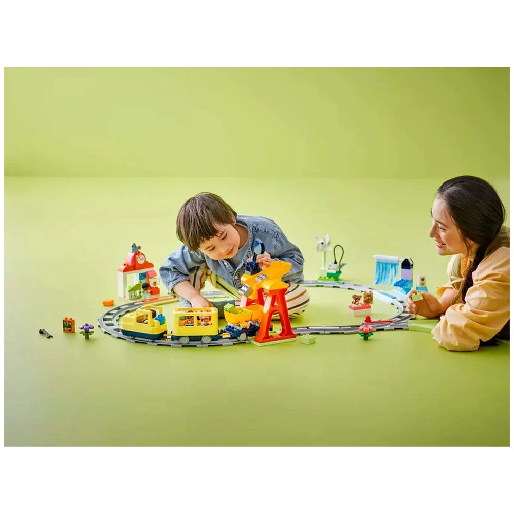 A child and a caregiver engage with the Lego Duplo Big Interactive Community Train set. The vibrant playset features a curved train track, colorful buildings, and figures, providing a fun learning environment. A joyful atmosphere is evident as they explore and create stories together on a bright green background.
