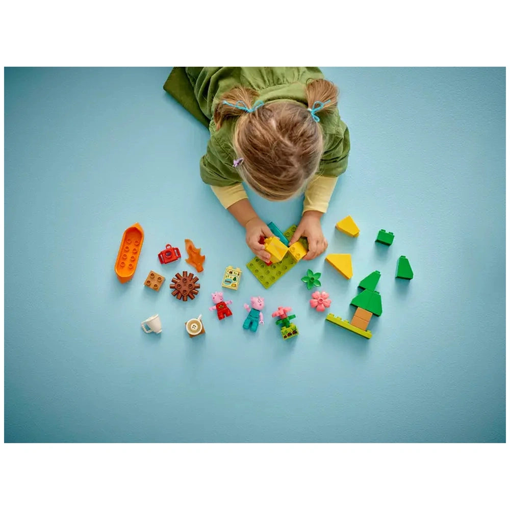 A child with blonde hair in pigtails plays on a blue surface, arranging colorful LEGO® DUPLO® pieces, including animals, trees, and camping accessories.