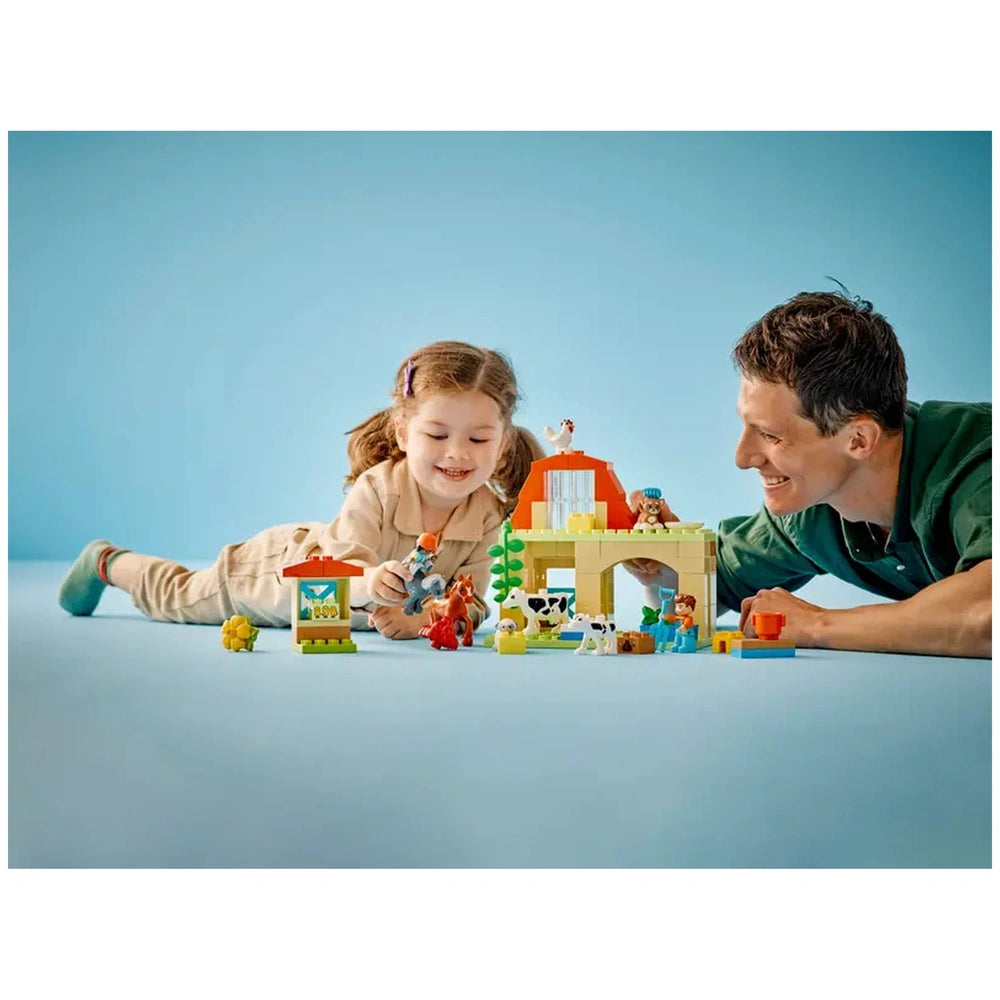 A young girl and a man play together with the LEGO® DUPLO® Caring for Animals at the Farm set, surrounded by colorful animal figures and a farmhouse, against a light blue background.