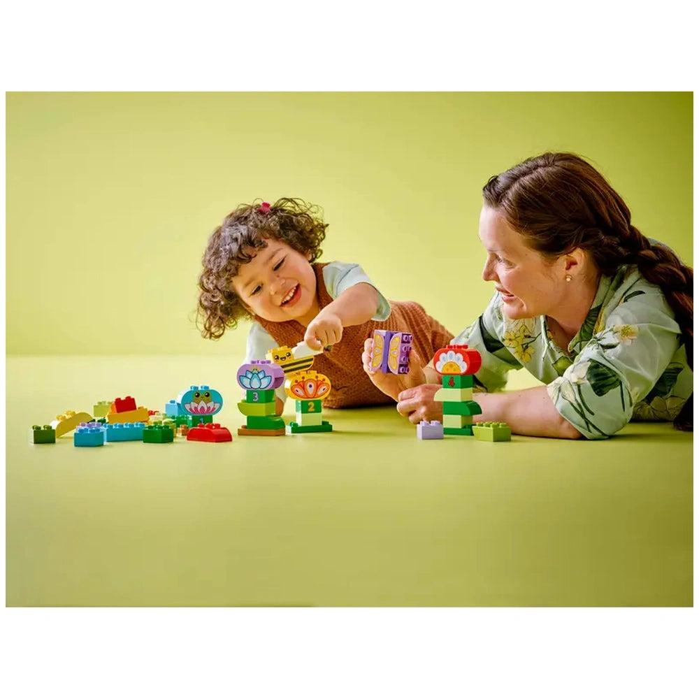 A young child and an adult enjoy playing with colorful LEGO® DUPLO® building blocks, constructing vibrant flowers and animals on a soft yellow surface.