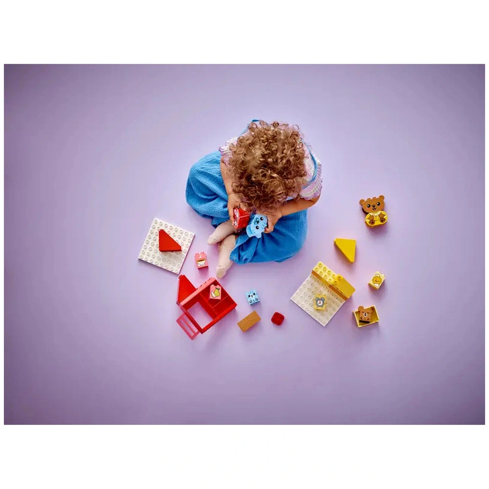 A toddler sits on a light purple surface, dressed in a blue outfit, playing with colorful LEGO® DUPLO® blocks. The child holds a blue animal figure and is surrounded by various pieces, including a red house, a yellow triangle, and animal figures, promoting imaginative play focused on daily routines.