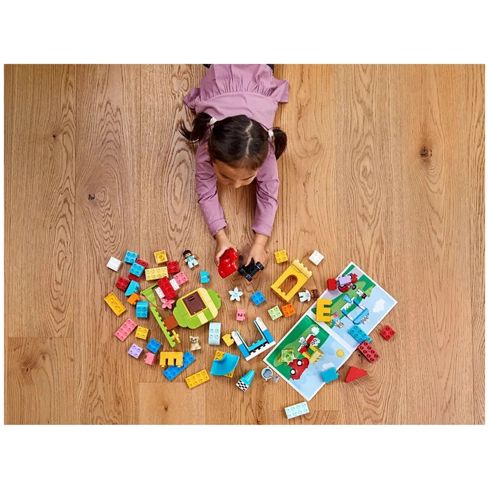 A child in a pink shirt plays with colorful LEGO® DUPLO® bricks on a wooden floor, surrounded by vibrant pieces and a building guide.