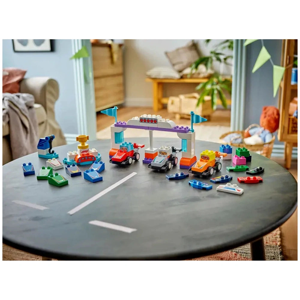 Colorful Lego Duplo F1® Team Race Cars set displayed on a black table, featuring multiple buildable race cars, a winner's podium, and racing accessories in a bright room.