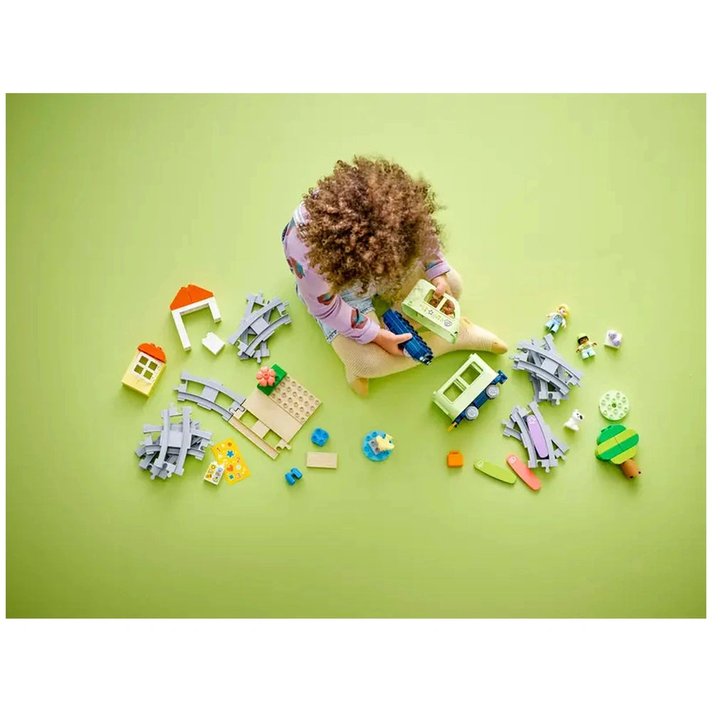 A child with curly hair sits on a green mat, focused on assembling the LEGO® DUPLO® Interactive Adventure Train. Surrounding them are colorful building bricks, train tracks, and small structures, including houses and trees, encouraging imaginative play.