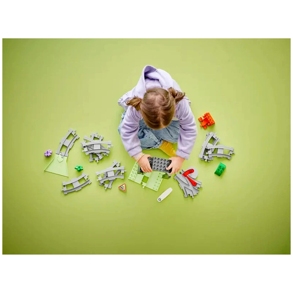 A young child in a purple hoodie sits on a green surface, assembling LEGO® DUPLO® train tracks and components, surrounded by colorful pieces, including a green triangle and red funnel.