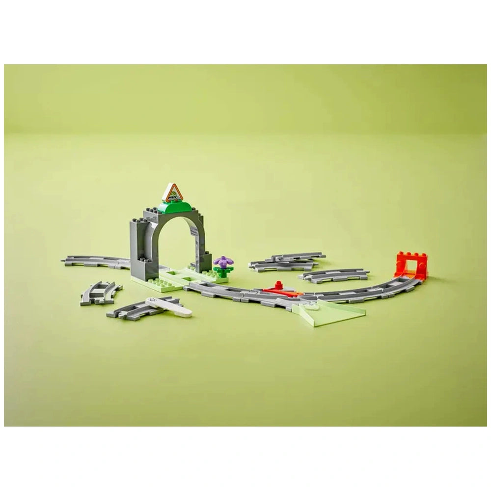 LEGO® DUPLO® Train Tunnel and Tracks Expansion Set featuring a gray archway with a green peak, colorful track pieces, and playful elements on a light green background.