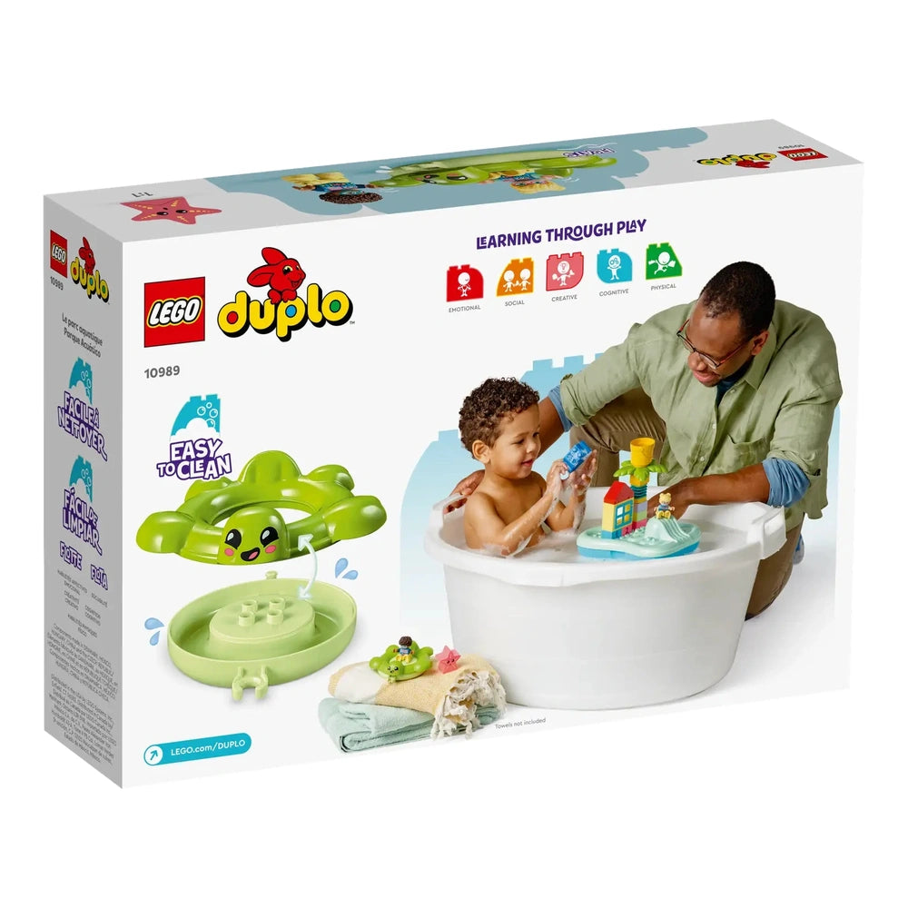 A toddler bathes happily in a white tub, playing with a colorful LEGO® DUPLO® Water Park set featuring a turtle floatie and a palm tree, while an adult interacts with him. Towels are neatly arranged nearby.