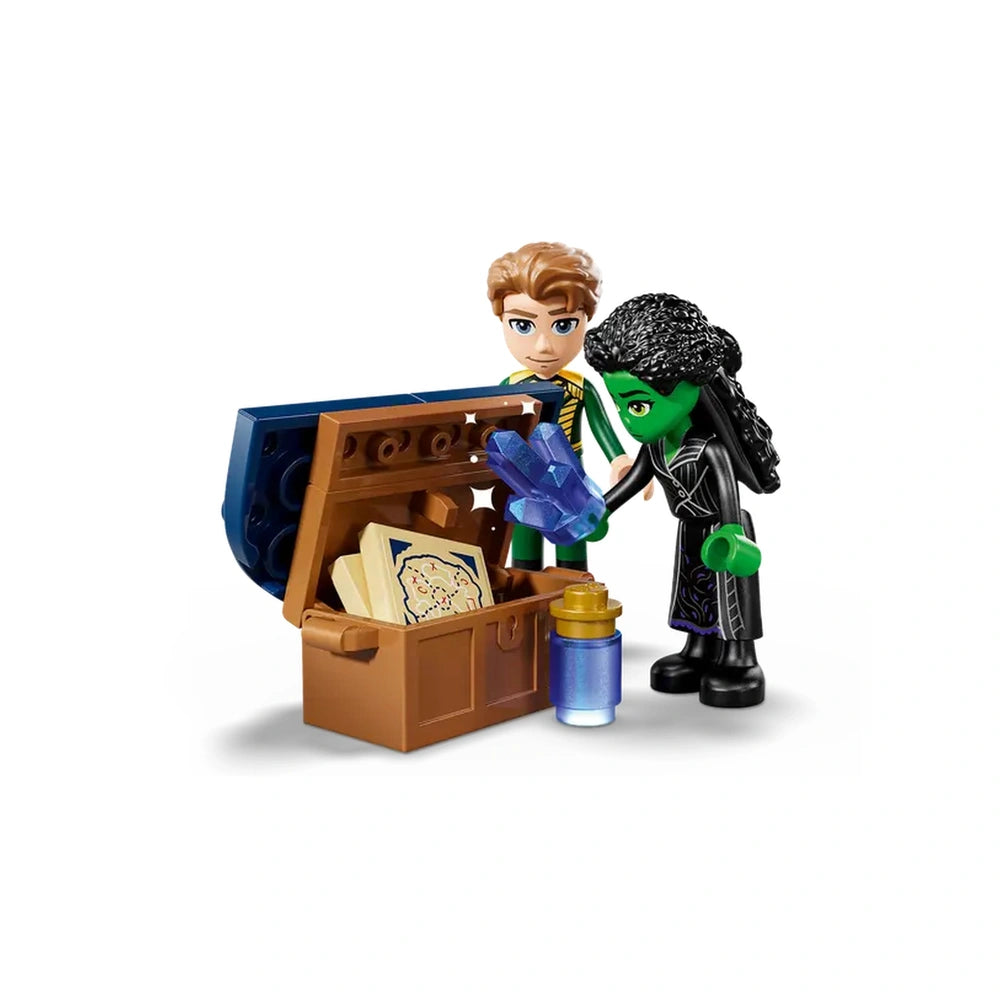 Elphaba, a green-skinned figure in a black dress, holds a blue crystal while standing next to a blonde character, both peering into a treasure chest. The chest is open, revealing a book and a small jar alongside the crystal.