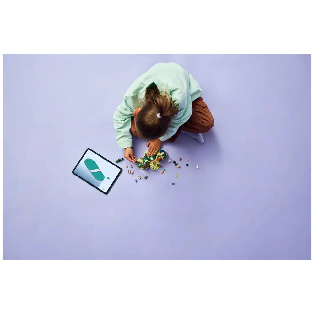 A child in a light green sweater sits on a soft purple surface, assembling LEGO pieces into a whimsical design, with a tablet displaying instructions nearby.
