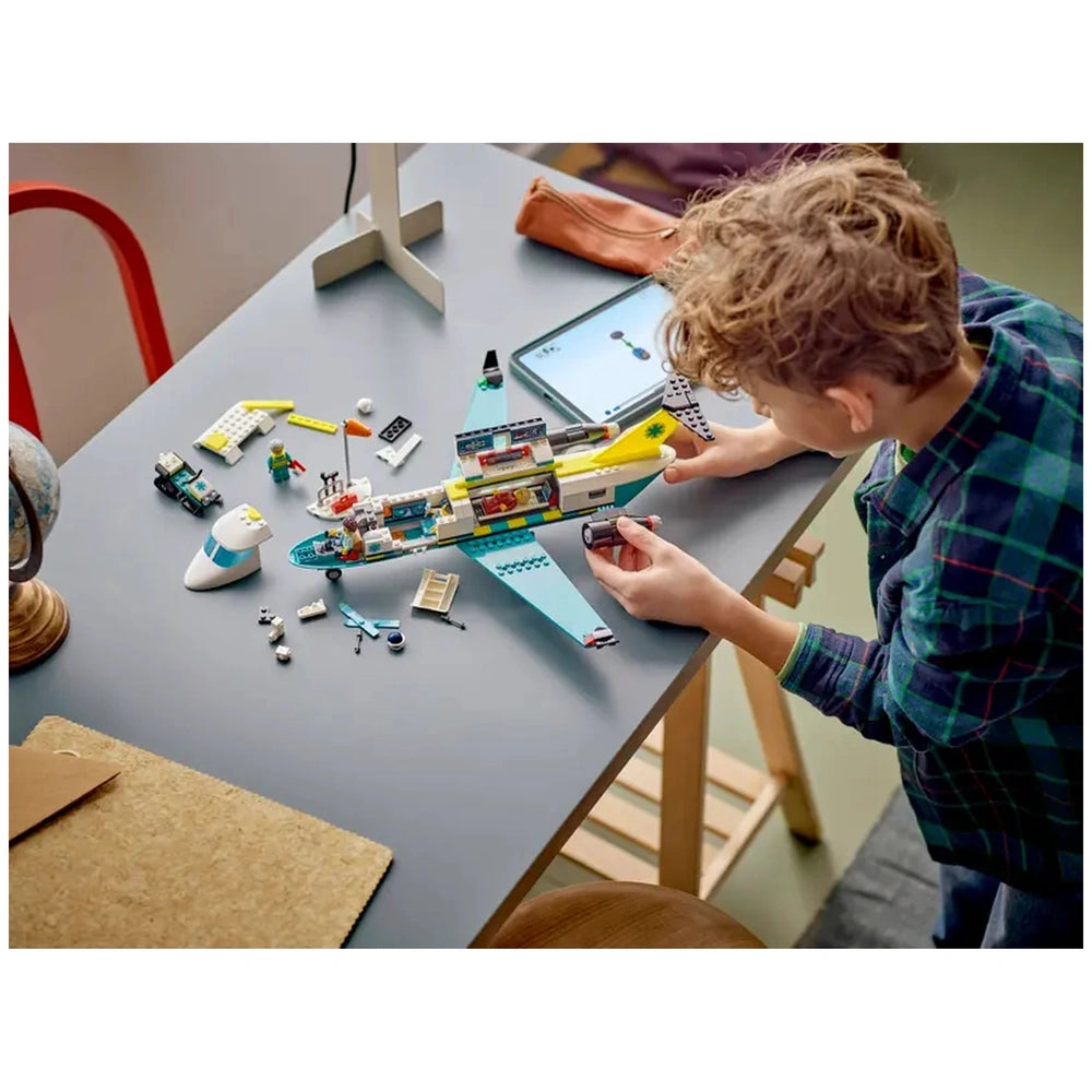A child with curly hair builds a colorful LEGO emergency air ambulance plane on a gray table, surrounded by additional pieces and minifigures.