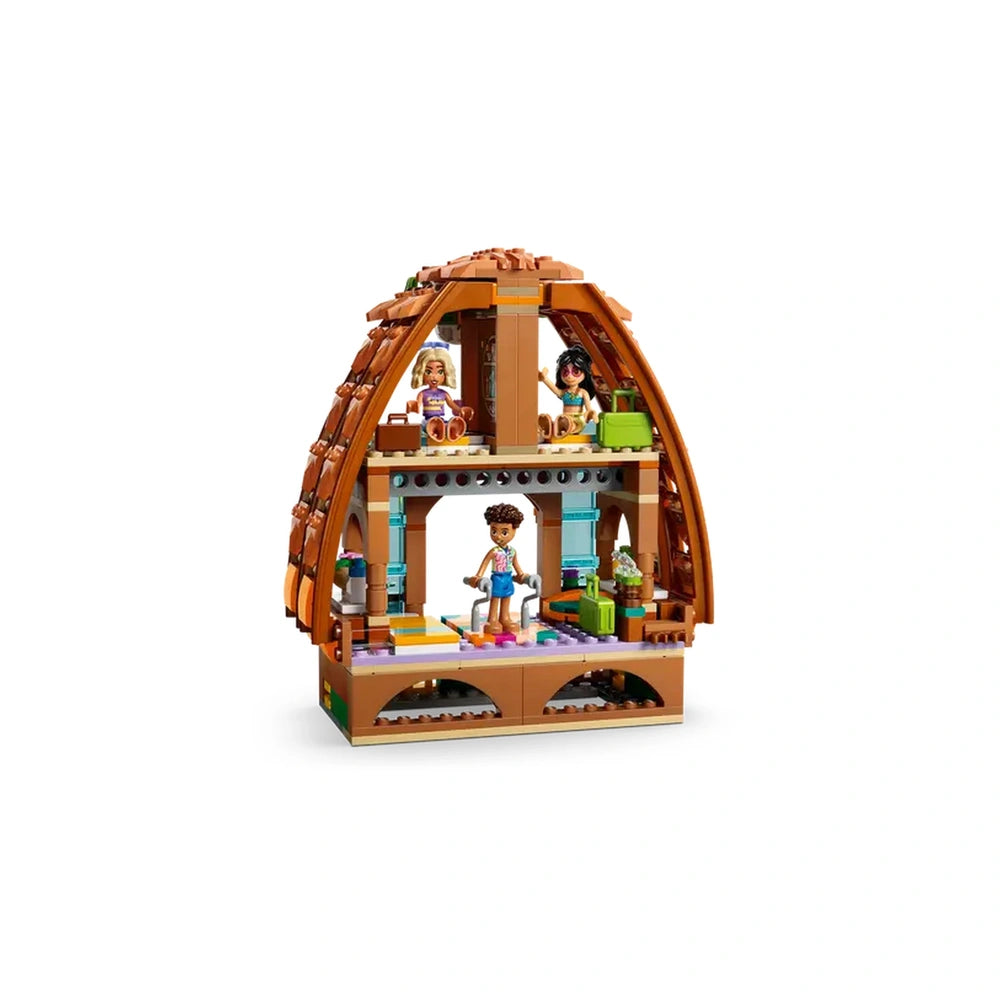 Three LEGO Friends minidolls are positioned inside a colorful beach resort set, with two sitting on a raised area and one standing playfully. The interior features a vibrant design, showcasing beach-themed decor and activities. Various accessories, including a small plant and furniture pieces, enhance the scene, creating an inviting atmosphere for imaginative play. The structure has a distinctive brown, wave-like roof, giving it a unique beach house aesthetic.