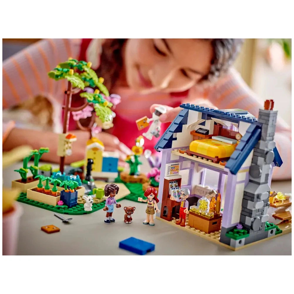 A child is joyfully playing with the LEGO Friends Beekeepers’ House and Flower Garden set, featuring a charming two-story house and a vibrant garden. Two minidolls are visible alongside a small dog and a cat, surrounded by colorful plants and accessories. The scene captures a moment of creativity and imagination as the child engages with the intricate details of the set.