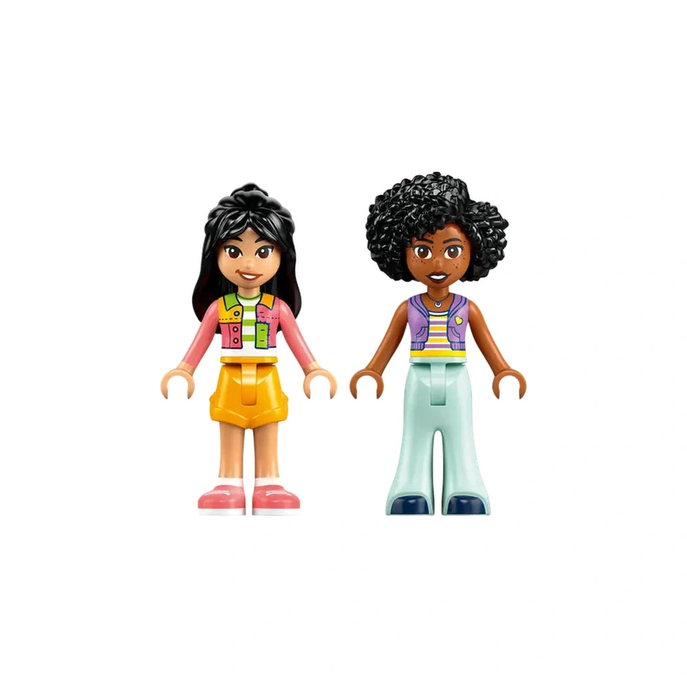 Two LEGO Friends minidolls stand side by side. On the left, a girl with black hair in a pink vest and yellow shorts smiles, while on the right, a girl with curly black hair wears a multicolored top and light blue pants. Both have cheerful expressions, showcasing vibrant colors and playful designs that encourage imaginative play.