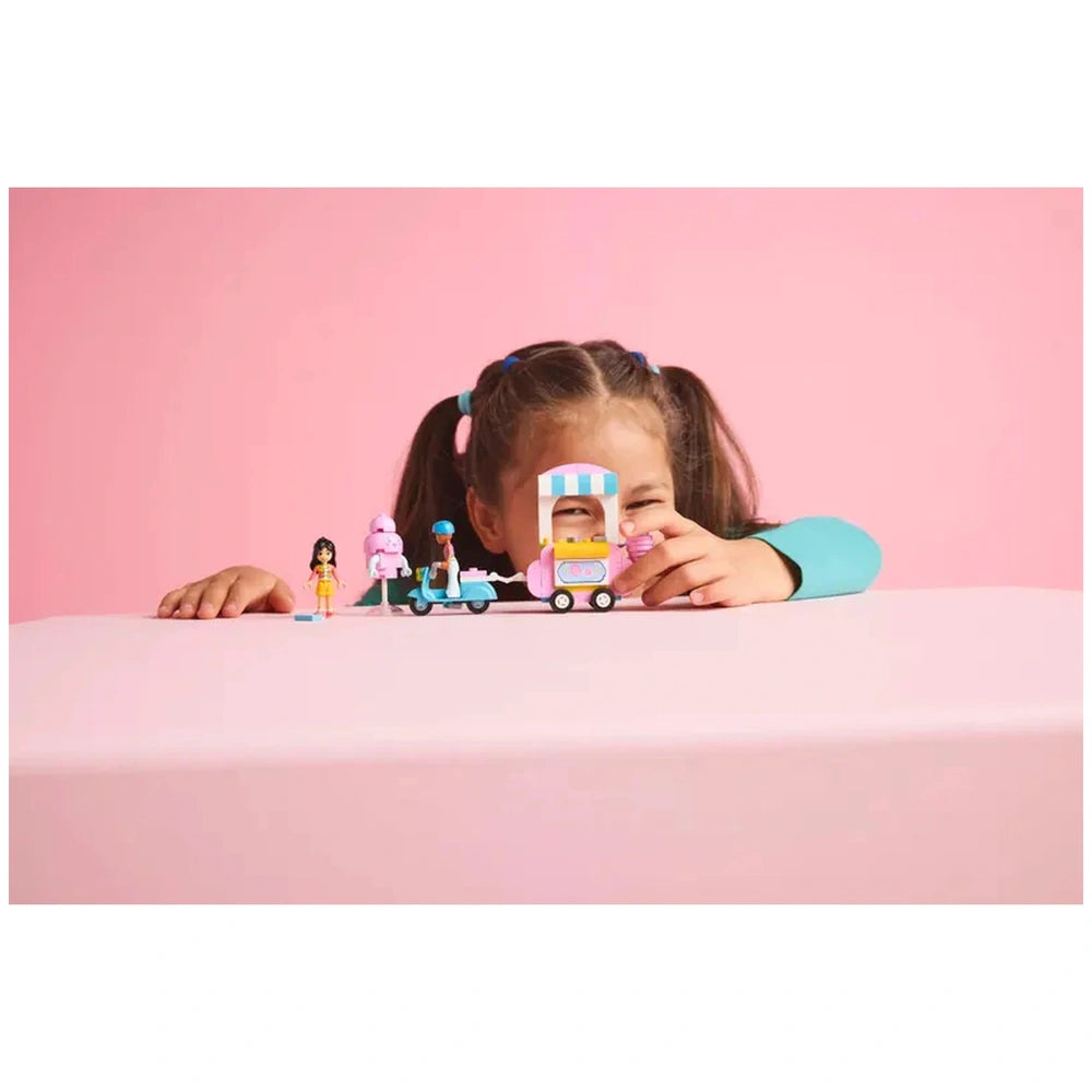 A smiling child with brown hair and pigtails plays with the LEGO® Friends Cotton Candy Stand and Scooter set, featuring two minidolls and a pastel-colored food cart against a pink background.