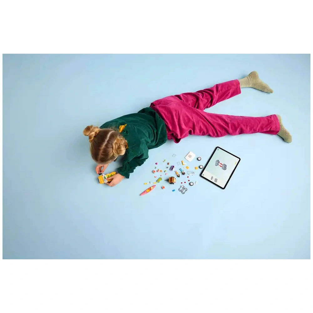 A child with blonde hair and pink pants is lying on a blue surface, building a LEGO Friends travel car. Various colorful LEGO pieces are scattered around, alongside a tablet displaying a character.
