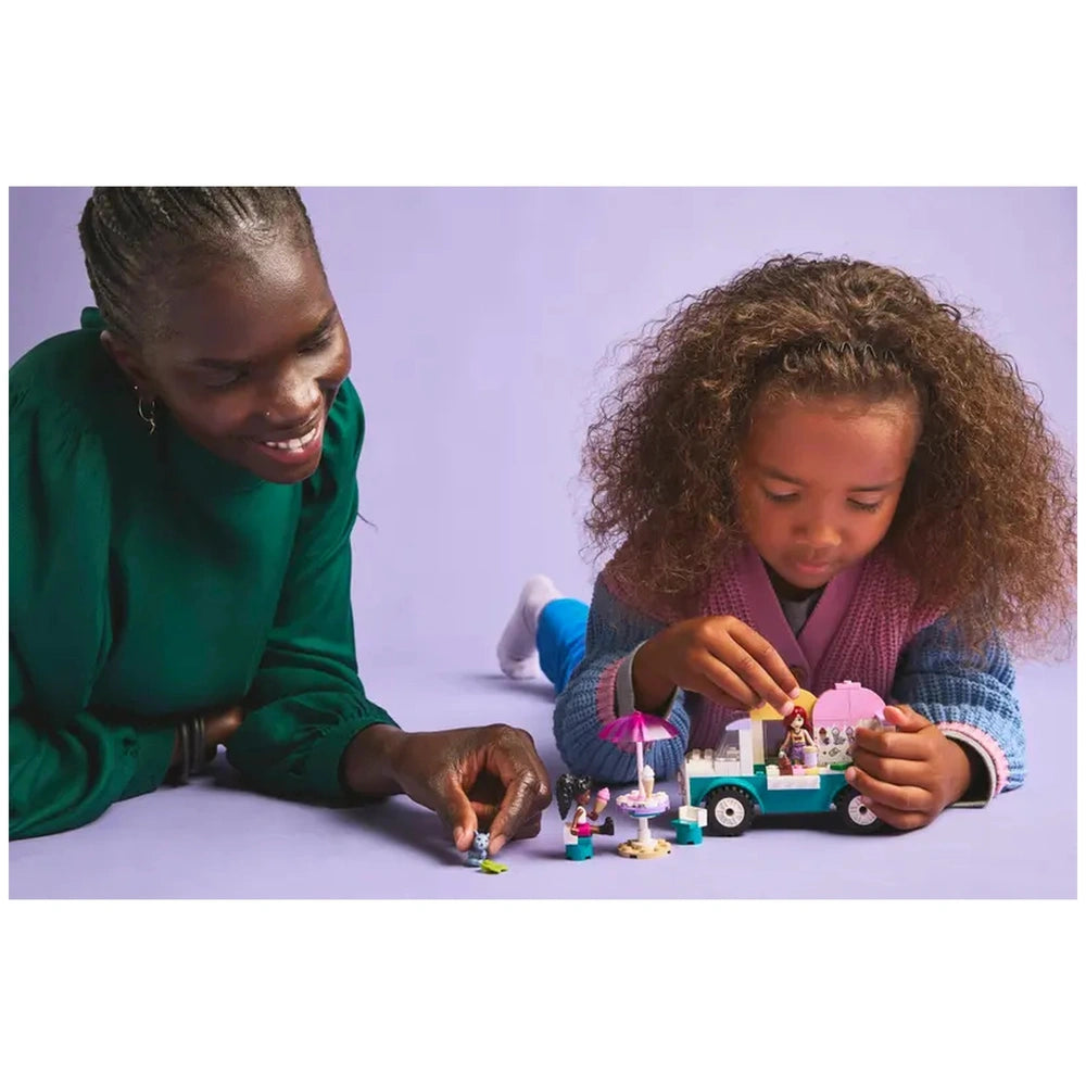 A child playing with the LEGO® Friends Heartlake City Ice Cream Truck set, surrounded by colorful toy figures and accessories, while an adult smiles nearby.