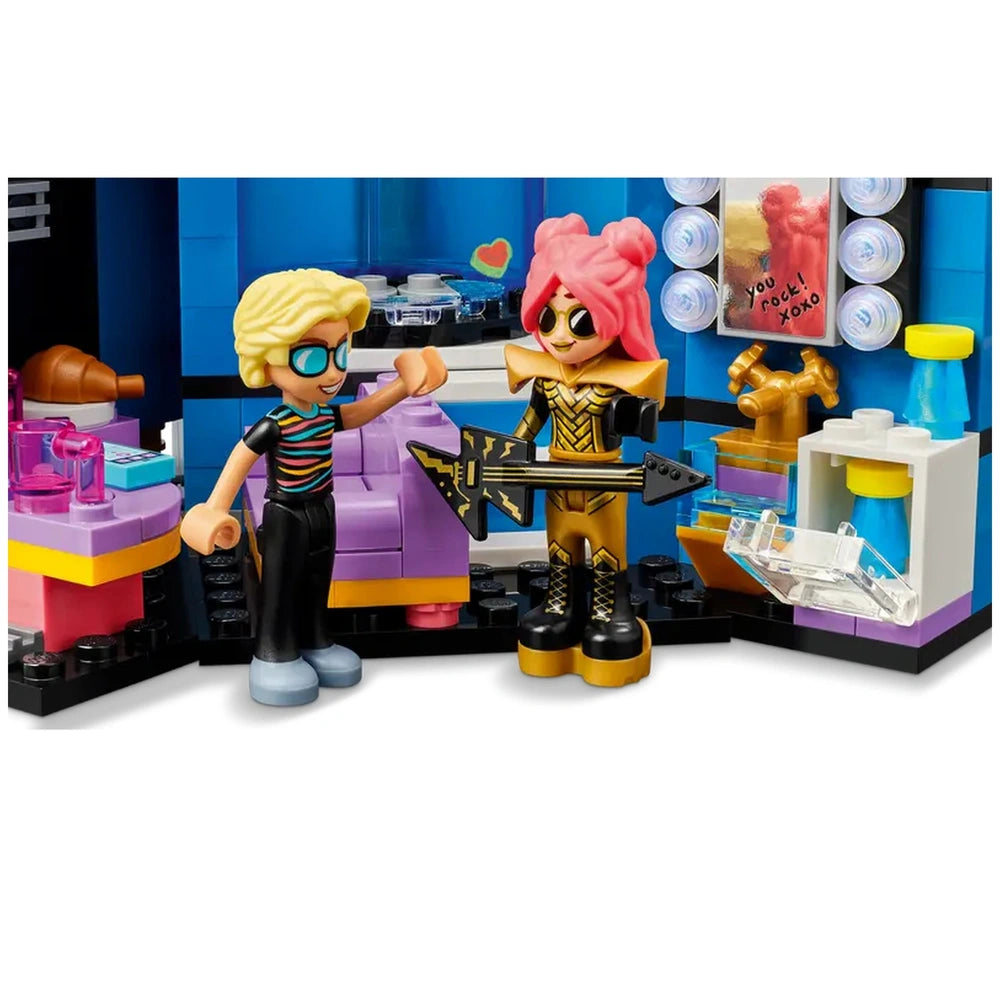 Two colorful mini-dolls in a music-themed playset, one holding a guitar and the other gesturing excitedly. Bright pink and gold outfits contrast with a purple stage backdrop featuring musical accessories.