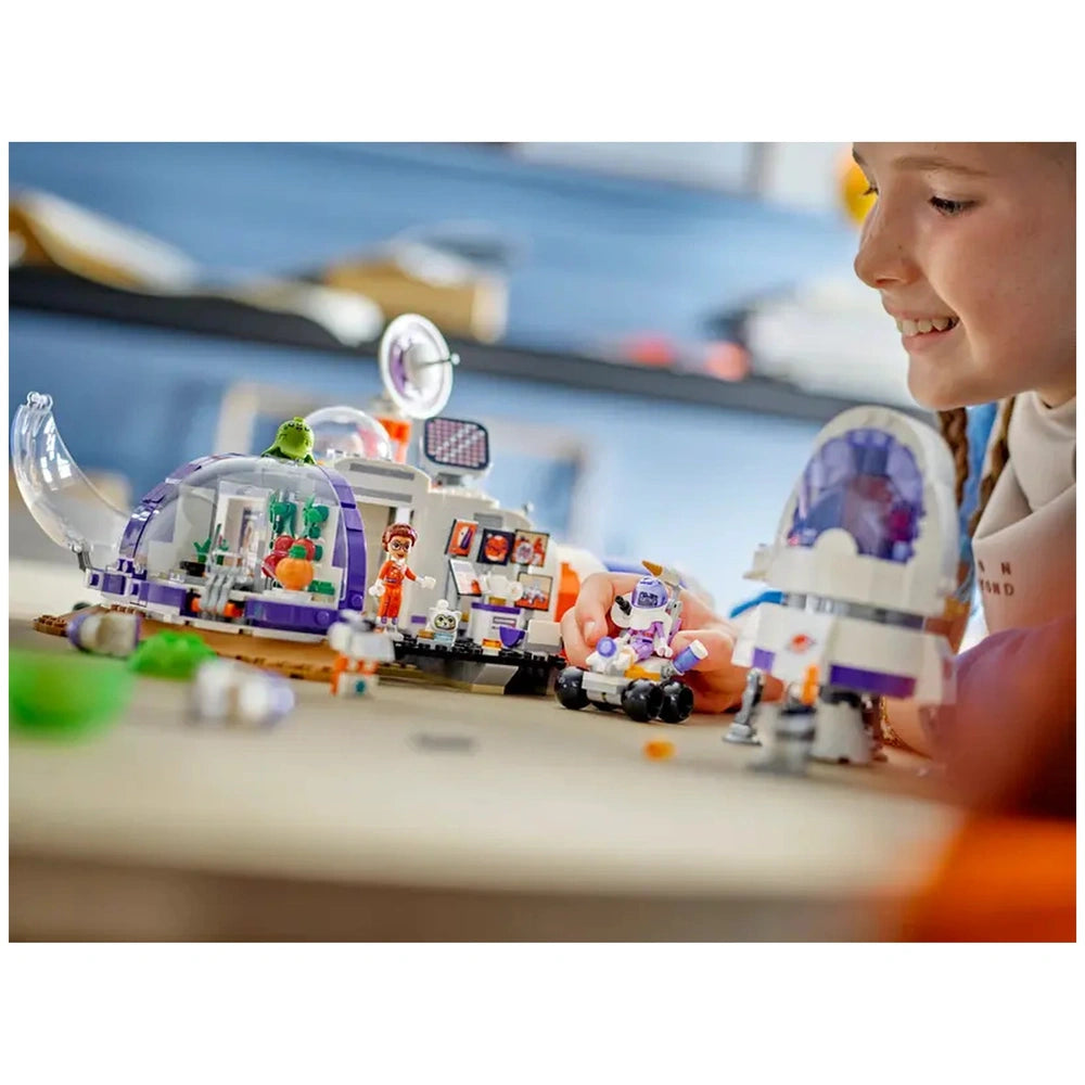 A young girl smiles while playing with the LEGO® Friends Mars Space Base and Rocket set. The colorful modular base features a transparent dome, mini-dolls, and a rover. Various space-themed accessories lay scattered around, inviting imaginative exploration.